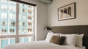 A hotel room with a large window, a bed with white bedding, a brown pillow, a black lamp, and a framed picture on the wall. - Luxury Designer Loft 302 | River North (Chicago)