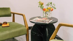 En grøn polstret stol med et par solbriller og en bog ved siden af. Et rundt, lille bord med en sort marmortop har et magasin og to klare vaser med blomster.