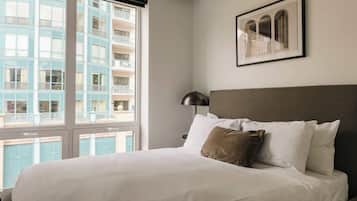 A hotel room with a large window, a bed with white bedding, a brown pillow, a black lamp, and a framed picture on the wall.