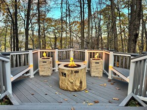 A deck with a central fire pit surrounded by stone planters, set against a backdrop of trees.