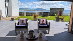 An outdoor seating area with two chairs and a table, a stone wall, and a view of the sea and hills.