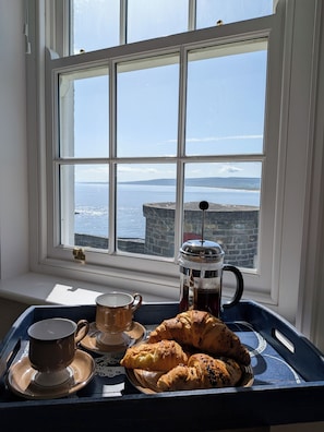 Un vassoio con una caffettiera, una tazza e dolci, vicino a una finestra vista mare.
