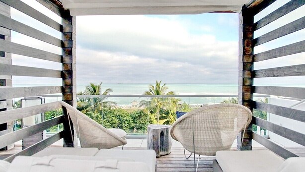 Een balkon met twee rieten stoelen en een witte bank, met uitzicht op een strand en de oceaan.