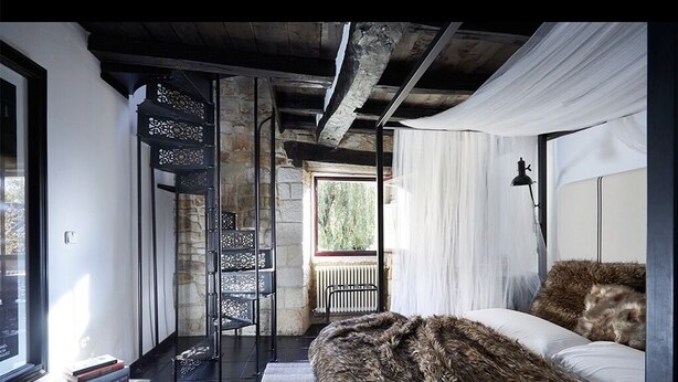 Une chambre avec une grande moquette en fourrure, un lit recouvert de draps blancs, une chaise en métal noir et une fenêtre donnant sur un paysage verdoyant.