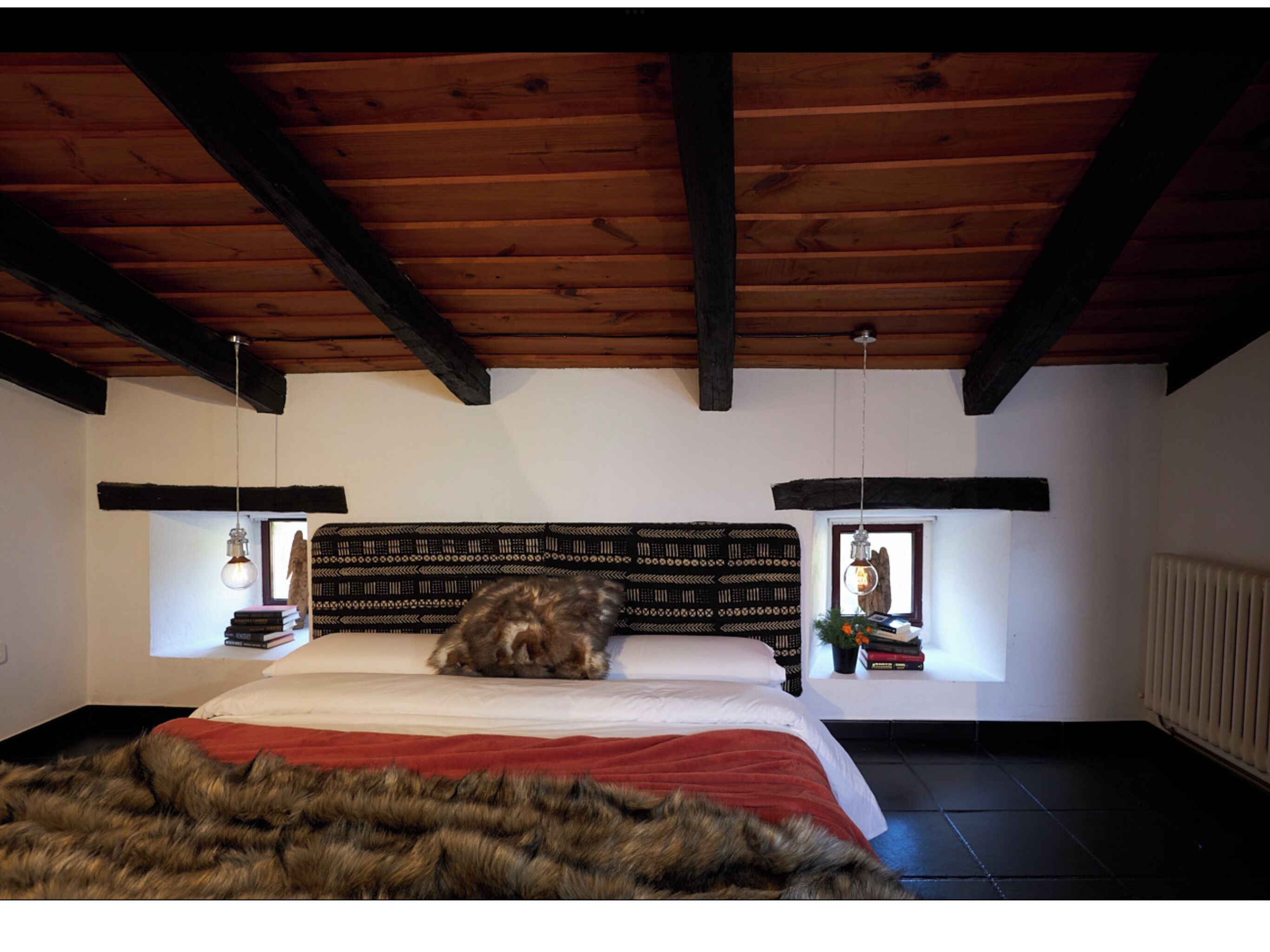 Une chambre à coucher avec un plafond en bois, une tête de lit à motifs, une couvre-lit rouge et deux tables de chevet ornées de livres et d’objets décoratifs.