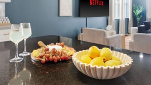 A modern living room with a flat-screen TV displaying 'NETFLIX', a fruit platter, and two glasses of white wine on a kitchen counter. - Golf Sim, Theater, Games, Walk to Beach, Sleeps 28 (North Myrtle Beach)