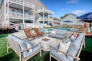 Un espace aménagé au bord de la piscine, avec des sièges d’extérieur, un foyer et un bâtiment à deux étages doté de balcons.