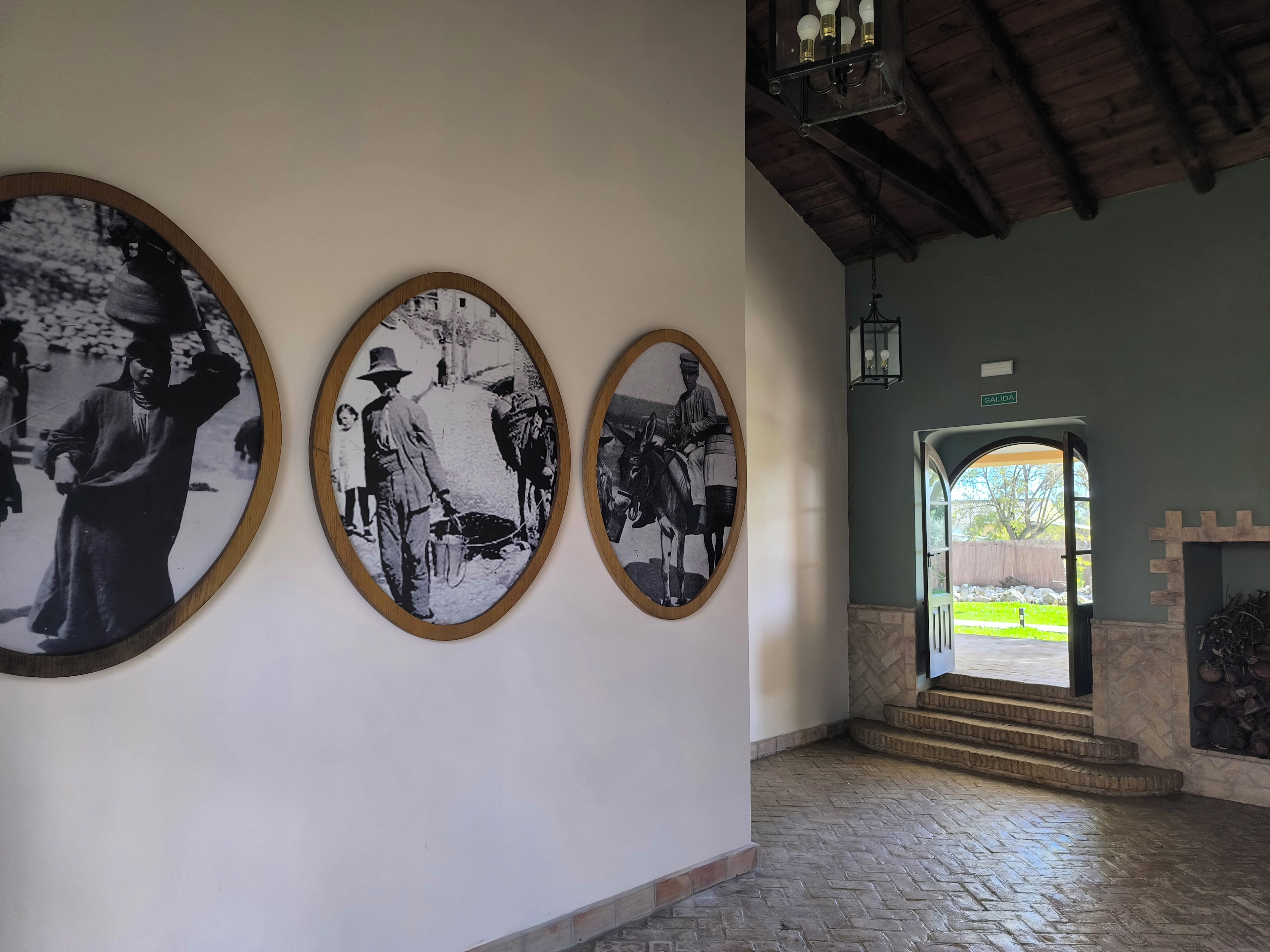 Un couloir avec trois photographies en noir et blanc encadrées accrochées au mur, un plafond en bois et une porte marquée du mot « SORTIE » qui donne sur l’extérieur.