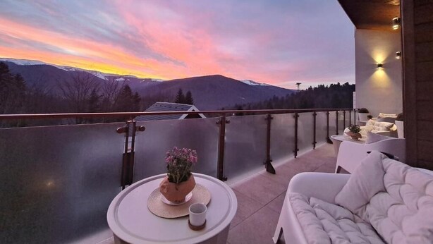 Un balcón con vista a las montañas al atardecer, un sofá blanco y una pequeña mesa con una planta y una taza.