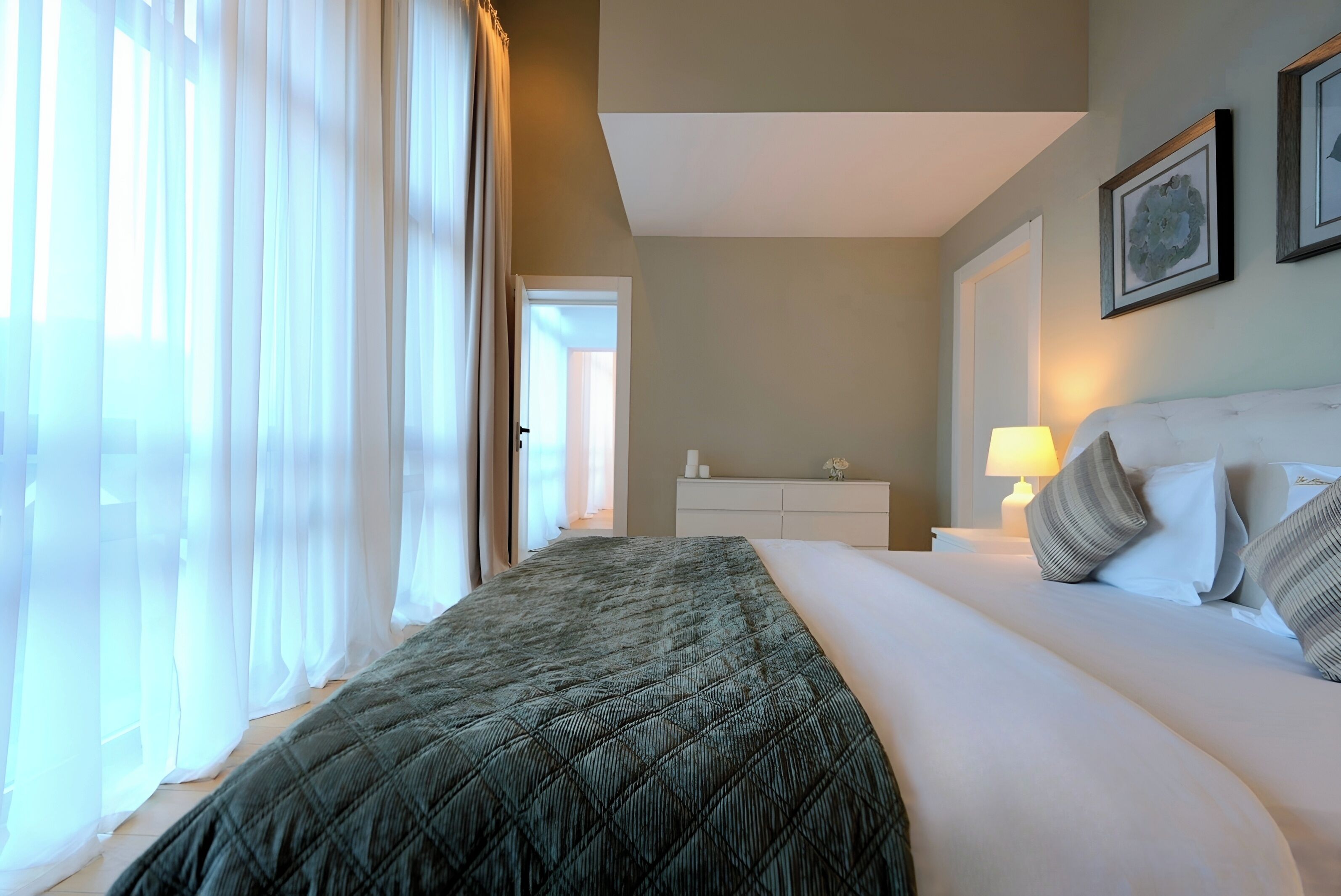Habitación de hotel con una cama grande, una mesita de noche con lámpara, y vistas al pasillo.