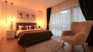 Un dormitorio con cama, un sillón mullido, una mesita de noche con una pequeña planta y una ventana con cortinas traslúcidas.