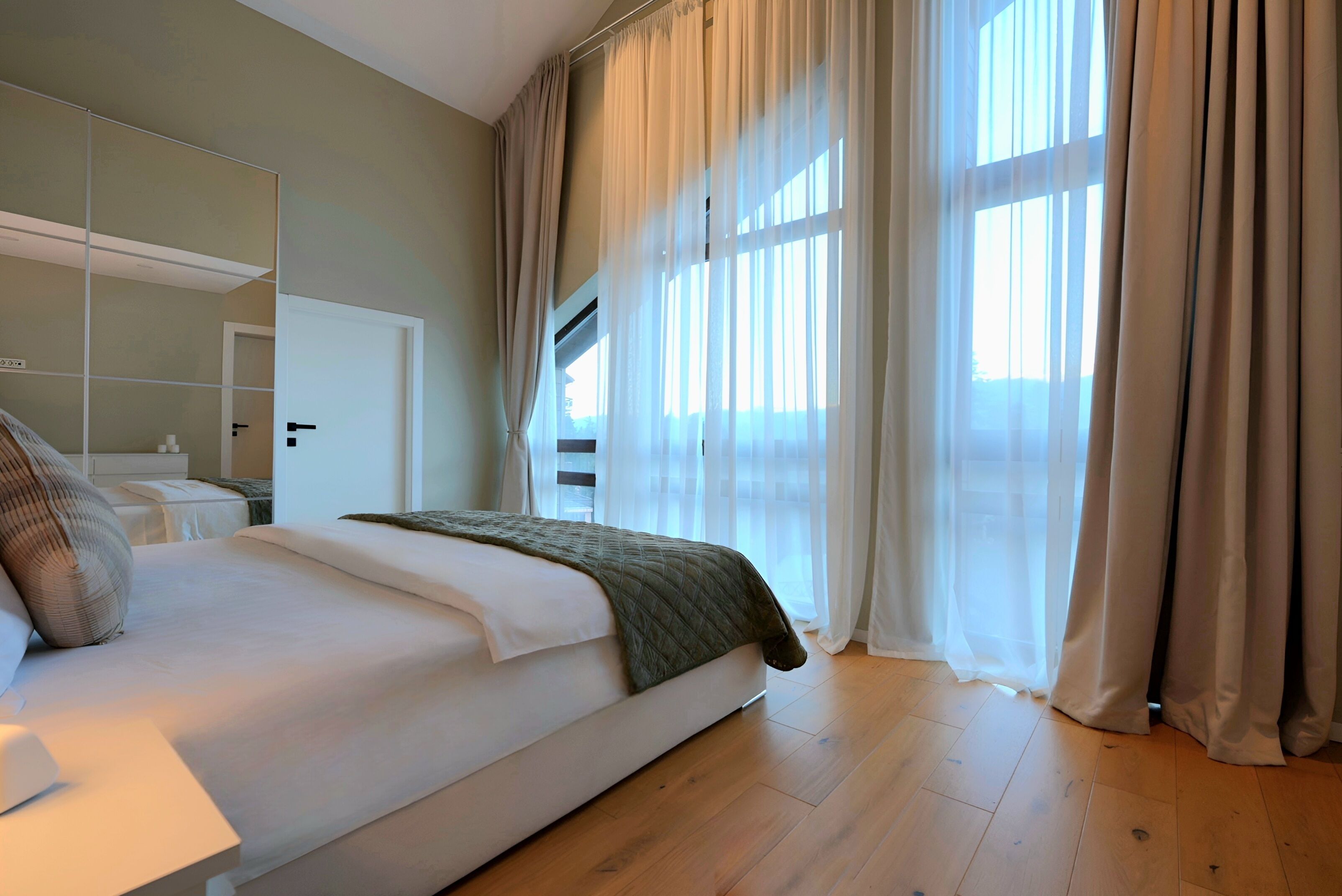 Un dormitorio moderno con una cama grande, una mesita de noche, un espejo y cortinas translúcidas.