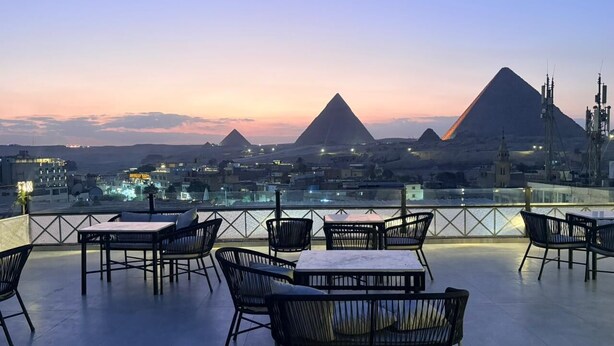 A rooftop terrace with tables and chairs, overlooking pyramids and a cityscape at dusk. - Pyramids Pavilion Boutique Hotel (Giza)
