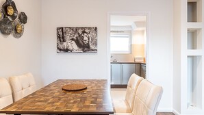 Un comedor con una mesa de madera y sillas acolchadas, un cuadro de un león blanco y negro en la pared, y vista a la cocina.