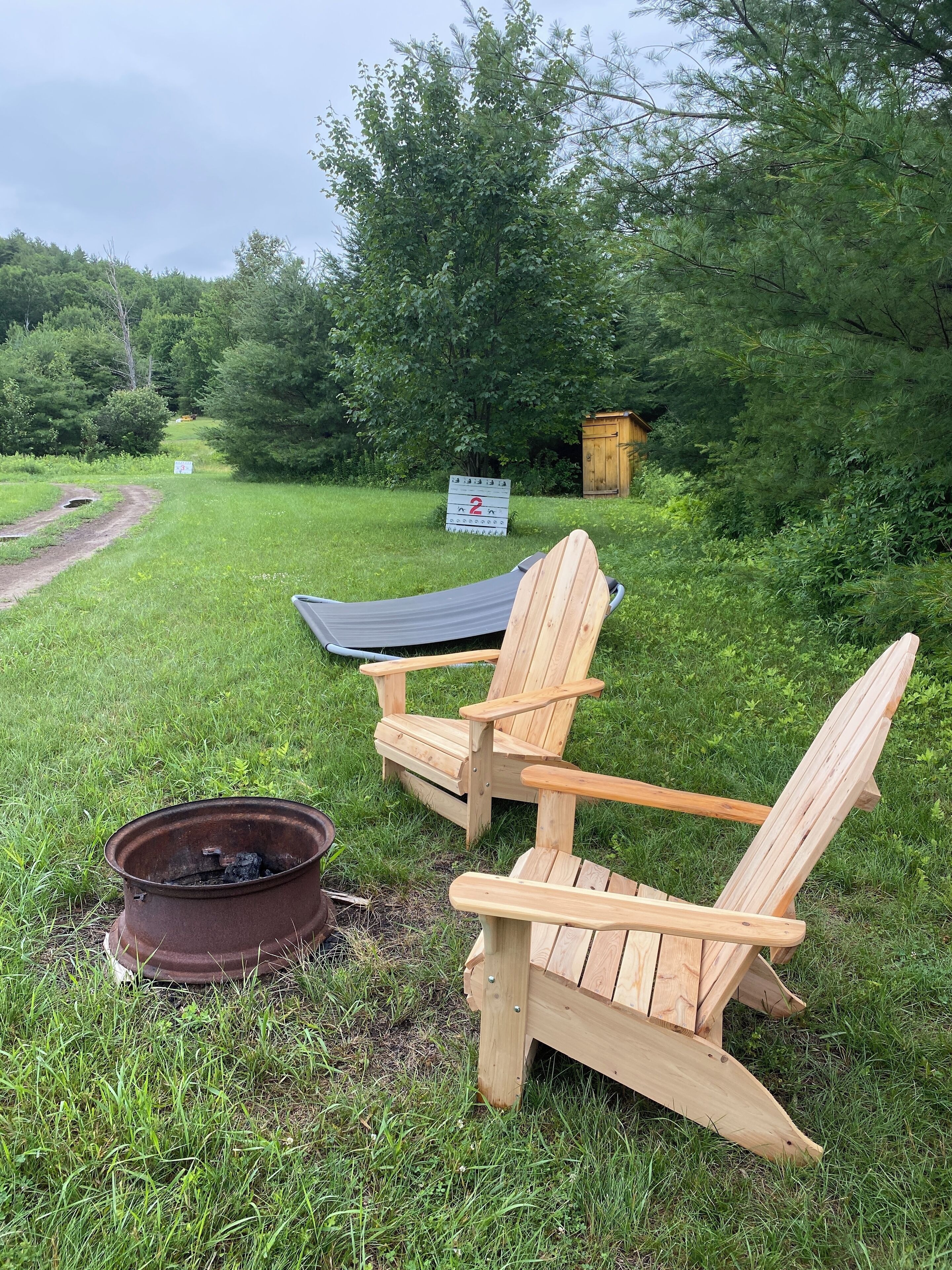 Zwei Holz-Adirondack-Stühle, eine Metall-Feuerschale und eine Holz-Aborteinrichtung in einer grasbewachsenen Fläche mit Bäumen im Hintergrund.