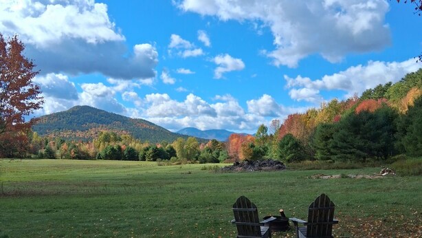 A picnic table with two Adirondack chairs in a grassy field with autumn foliage and a mountain in the background.