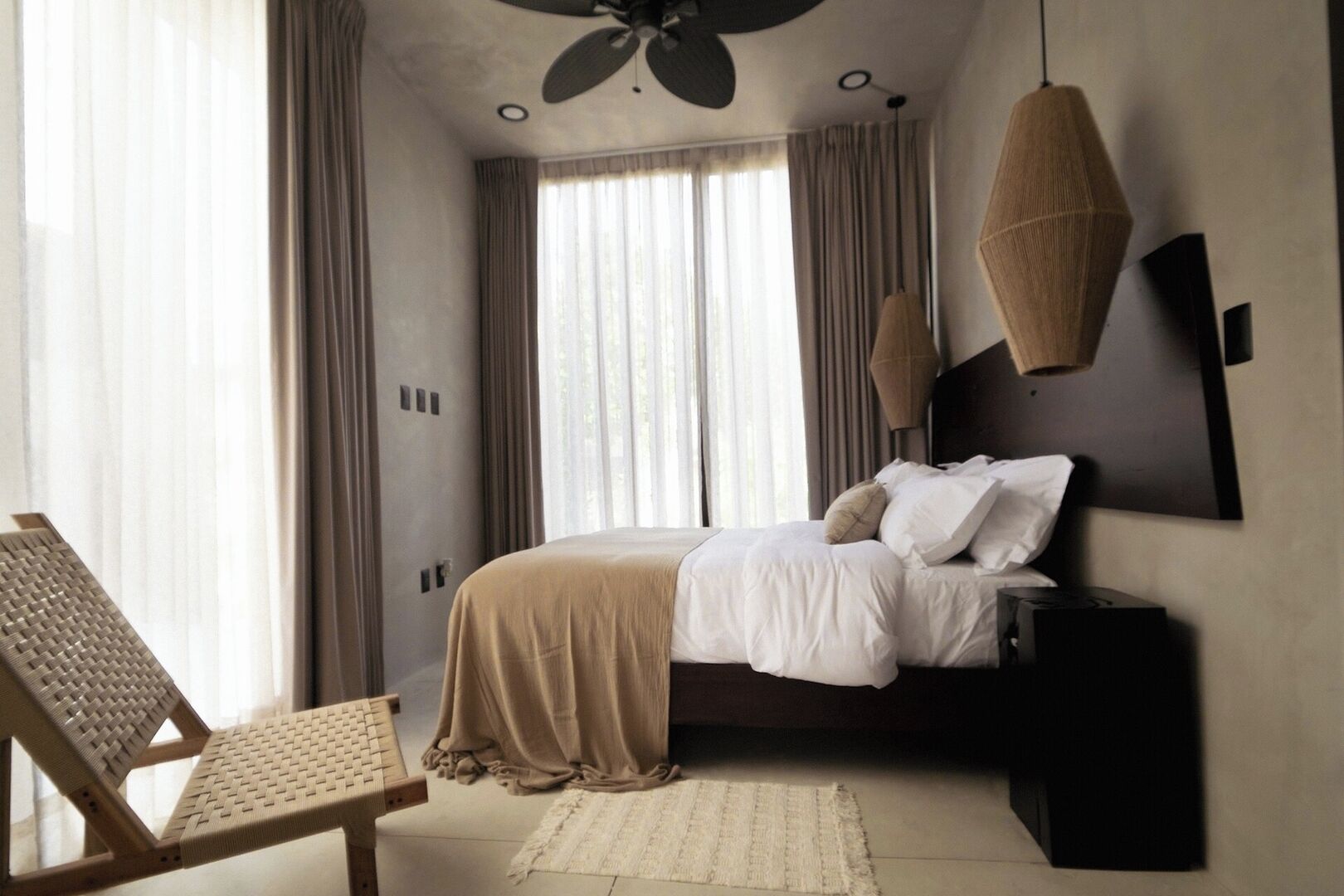 Un dormitorio con cama, una silla, un escritorio, ventilador de techo y un ventanal con cortinas.