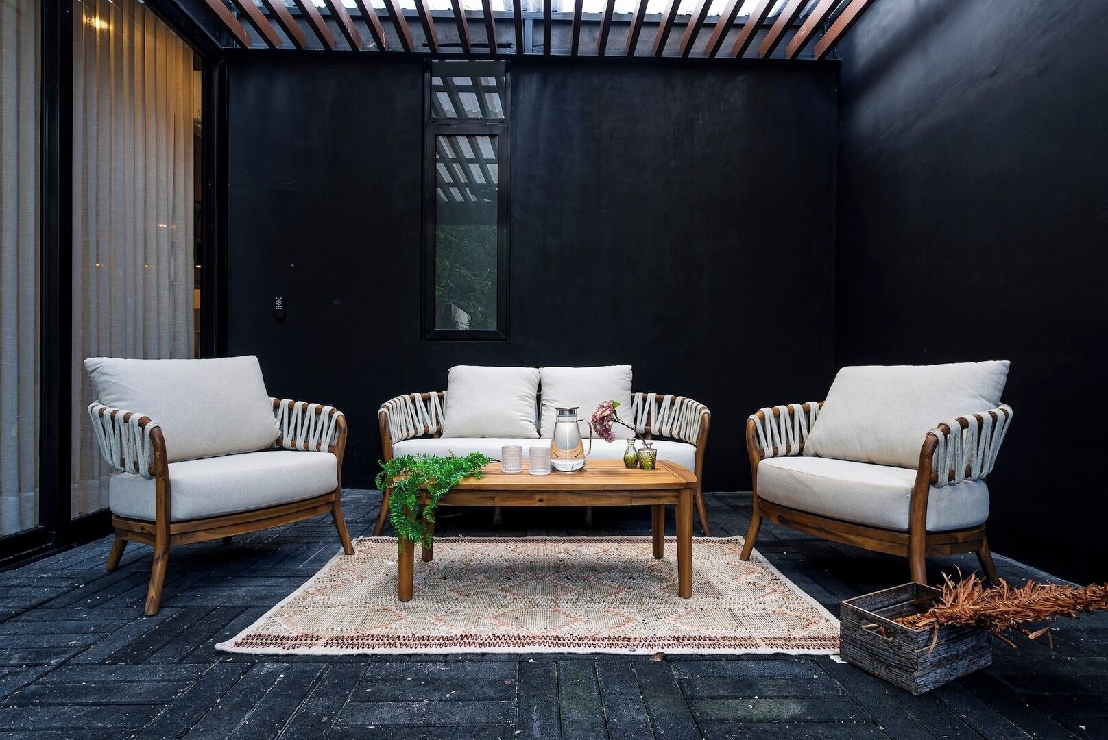 Uma área externa moderna com assento, incluindo duas cadeiras de vime e uma mesa de madeira, em frente a uma parede preta com uma janela.