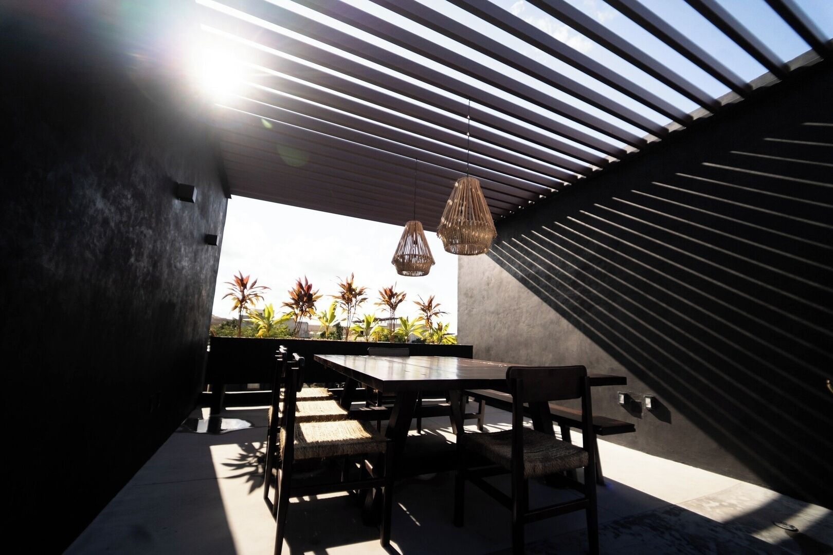 An outdoor dining area with a modern design, featuring a long table and chairs, a hanging light fixture, and a view of palm trees.