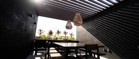 An outdoor dining area with a modern design, featuring a long table and chairs, a hanging light fixture, and a view of palm trees.