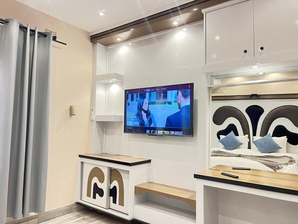 A modern living room with a flat-screen TV mounted on the wall, a white entertainment unit with a wooden top, and a grey curtain partially drawn. - Lindiwe House Self-Catering (Victoria Falls, Zimbabwe)