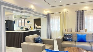 A modern living room with a sofa, armchairs, and a coffee table. There is a kitchen area with a refrigerator and a counter.