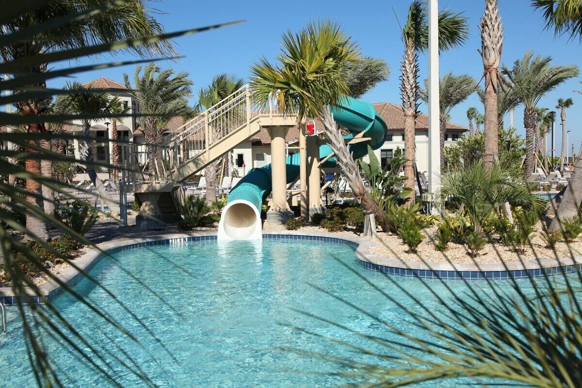 Ein Schwimmbecken mit Wasserrutsche, Palmen und ein Gebäude mit Balkon im Hintergrund.