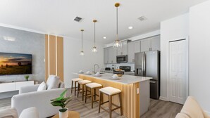 A modern kitchen with a central island, bar stools, and a dining area with a sofa and a TV. - Stylish Disney Getaway With Themed Rooms (Kissimmee)