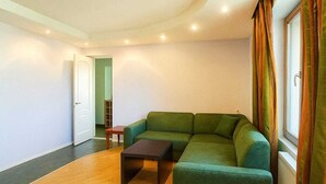 A modern living room with a green sectional sofa, a small wooden coffee table, and a view of a cityscape through a window with curtains.