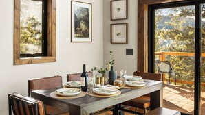 Un comedor con mesa de madera, sillas y un banco. Hay cuadros en la pared, una araña y un ventanal con vistas a árboles.