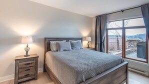 A bedroom with a wooden bed, two bedside tables with lamps, a large window with curtains, and a view of the outdoors. - Altitude Ski-in/Out at Resort with Private Hottub (Mont-Tremblant)