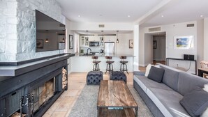 A modern living room with a stone fireplace, a grey sofa, a wooden coffee table, and a kitchen in the background. - Altitude Ski-in/Out at Resort with Private Hottub (Mont-Tremblant)