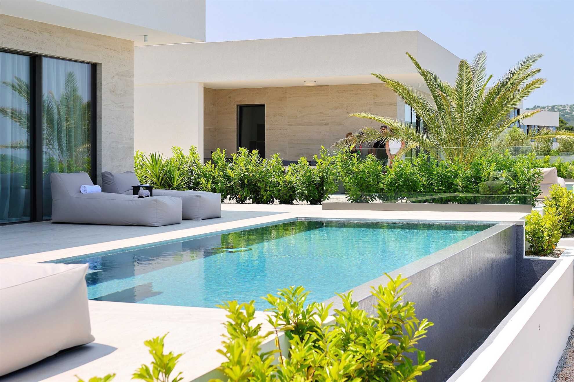 A modern outdoor pool area with a minimalist design, featuring a rectangular pool, a seating area with a sofa and chairs, and lush greenery.