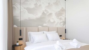 Una habitación de hotel moderna con una cama grande, ropa de cama blanca, un cabecero con arte mural con motivos de nubes, una mesita de noche con una planta y una mesita de noche con una lámpara.
