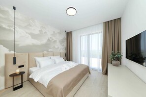 Une chambre moderne avec un grand lit, une table de chevet avec une bougie, un téléviseur fixé au mur et une grande fenêtre avec des rideaux.