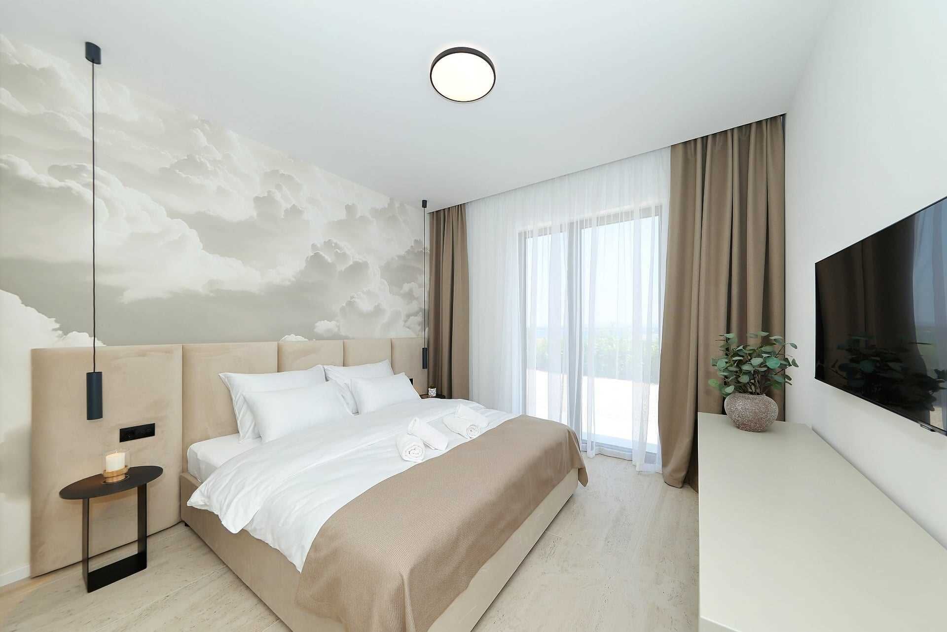 A modern bedroom with a large bed, a bedside table with a candle, a wall-mounted TV, and a large window with curtains.