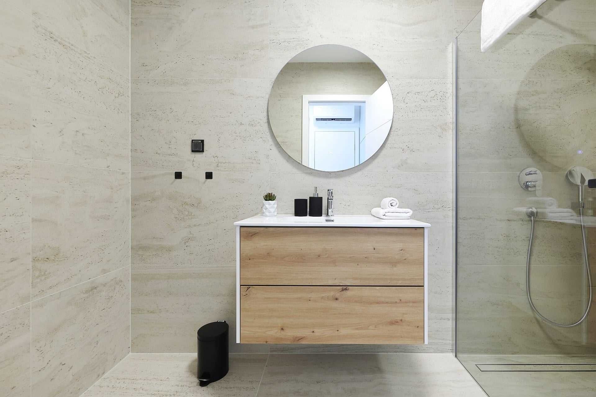 Ein Badezimmer mit rundem Spiegel, einem Holzwaschtisch, einem weißen Waschbecken und einer Duschkabine aus Glas.