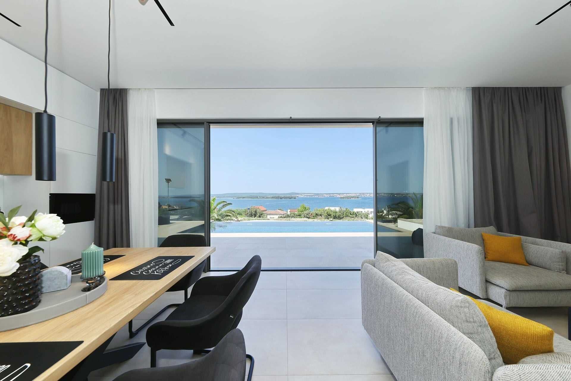 Un salon moderne avec vue sur la mer, un coin repas avec une table et des chaises, et un canapé agrémenté d’un coussin jaune.