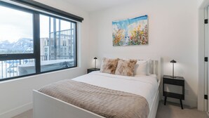 A bedroom with a large bed, two bedside tables, a window with a view of buildings and mountains, and a painting on the wall. - Avalanche Ridge (Revelstoke)