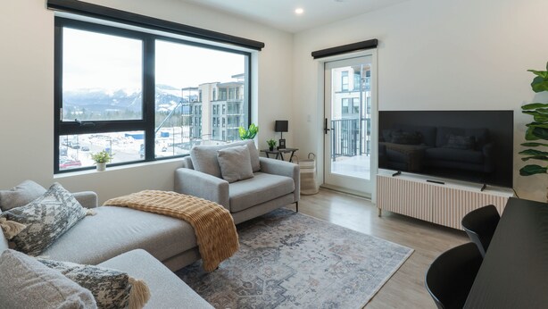 A modern living room with a sofa, a coffee table, a TV, and large windows offering a view of buildings and mountains. - Avalanche Ridge (Revelstoke)