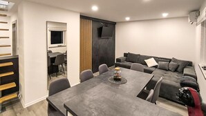 Ein modernes Wohnzimmer mit einem grauen Ecksofa, einem Esstisch mit Stühlen und einem an der Wand befestigten Fernseher.