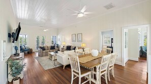 A spacious living area with a dining table, white chairs, a ceiling fan, and a television mounted on the wall. - Historic Retreat Near Beach w/ Pool 306 Charleston Blvd Roosevelt Retreat by Avantstay (Isle of Palms)