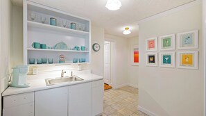 A kitchen with white cabinets, a sink, and a countertop with a Keurig coffee maker. There are shelves above the sink with glassware and teal mugs. A clock is on the wall, and framed pictures of cocktails are displayed on the adjacent wall. - Historic Retreat Near Beach w/ Pool 306 Charleston Blvd Roosevelt Retreat by Avantstay (Isle of Palms)