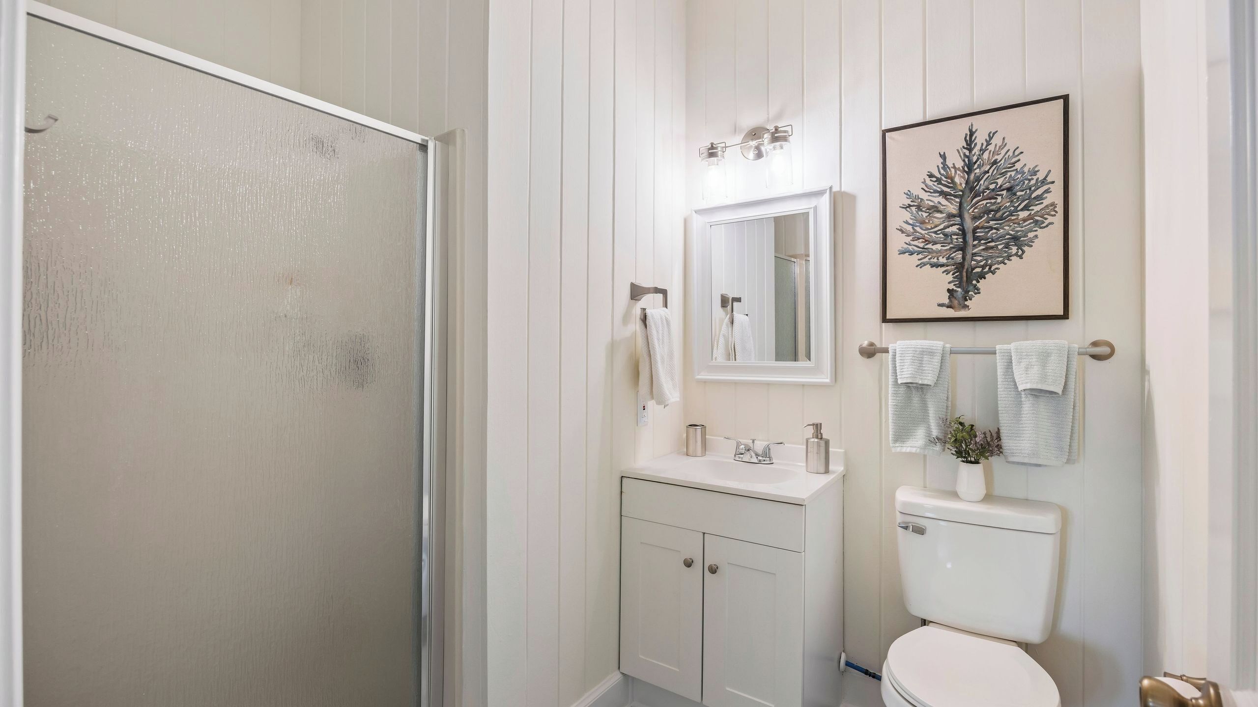 A bathroom with a shower, a sink with a mirror, a toilet, and a framed picture on the wall.