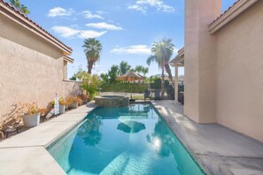 Ein rechteckiger Pool mit einer Wand aus Steinverkleidung, umgeben von einer Betonplattform, Palmen und einem überdachten Patio.