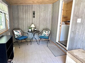 Ein Zimmer mit einer Holzwand, einem kleinen Tisch mit Lampe, zwei mit Kissen ausgestatteten Stühlen, einem fenster mit Vorhängen und einer Tür zu einem anderen Raum.