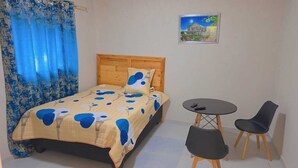 Een slaapkamer met een bed, een klein tafeltje, twee stoelen, een houten hoofdbord, een blauw gordijn en een ingelijst schilderij aan de muur.