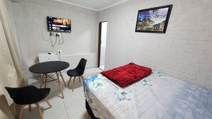 Een kleine, schone kamer met een bed, een bureau, een stoel en een aan de muur bevestigde televisie.