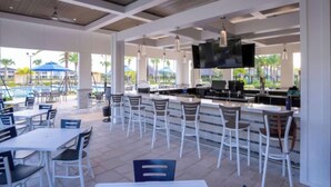 Un espace extérieur aménagé pour les repas, avec des tables et des chaises blanches, un bar doté d’un téléviseur à écran plat, et une piscine en arrière-plan.