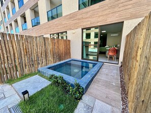 Egy modern, szabadtéri úszómedence, melynek köré fa kerítés van, egy kis kerttel és egy nagyméretű ablakokkal rendelkező épületre néz.